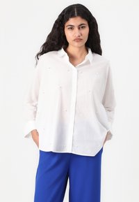 Chemise blanche à boutonnière fabriquée en tissu léger, avec des embellissements texturés. Assortie avec un pantalon bleu, elle a une coupe décontractée et des manches longues.