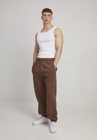 Débardeur blanc à côtes, pantalon de survêtement marron avec taille élastique et poches, et baskets blanches. Le modèle se tient devant un fond neutre.