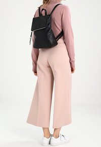 Czarny skórzany plecak z przednią klapą, srebrnym suwakiem i regulowanymi paskami. Noszony z bladoróżowymi szerokimi spodniami i białymi trampkami.