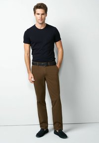 Zwart aansluitend T-shirt, bruin geruite broek, zwarte geweven riem en zwarte instapschoenen. Model staat tegen een lichte achtergrond.