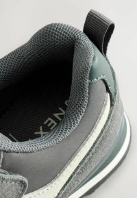 Nahaufnahme des gepolsterten Kragens eines grauen Sneakers, der Zuglasche, der genähten Wildleder- und Stoffabschnitte sowie eines Teils der strukturierten Innensohle mit teilweise sichtbarem Text.
