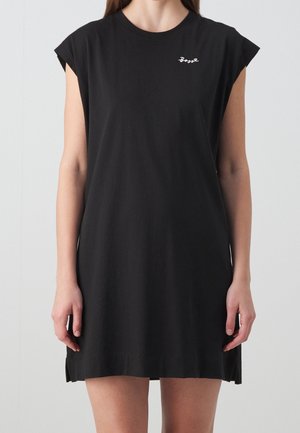Femme portant une robe noire sans manches avec le texte en écriture cursive blanche « Boys » sur la partie supérieure gauche de la poitrine, sur un fond clair uni.