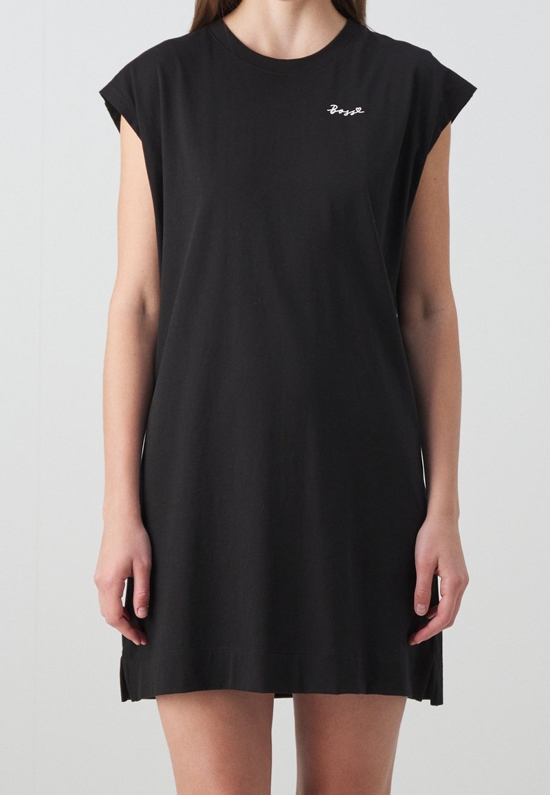 Femme portant une robe noire sans manches avec le texte en écriture cursive blanche « Boys » sur la partie supérieure gauche de la poitrine, sur un fond clair uni.