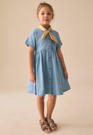 Young girl wearing a light blue button-front dress, yellow patterned neck scarf, and leopard-print sandals, standing on a beige background.