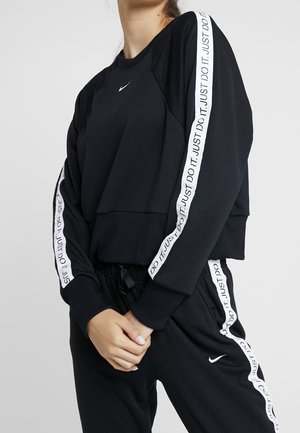 Kobieta ubrana w czarny dres Nike z białymi paskami „Just Do It.” na rękawach i spodniach, ręce złączone z przodu, głowa częściowo poza kadrem.