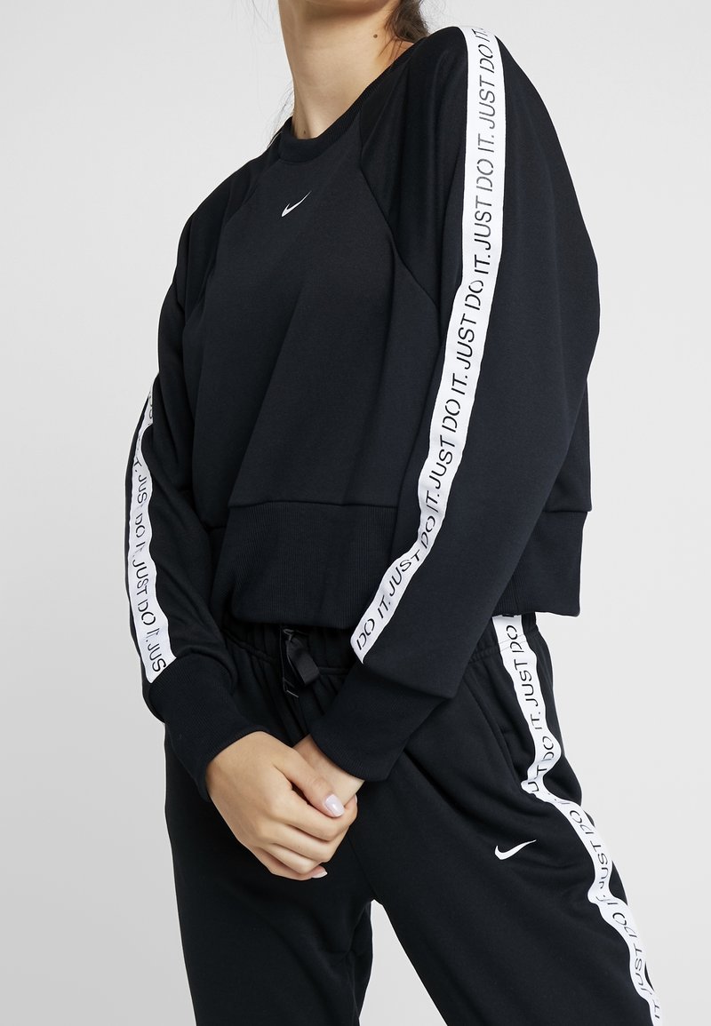Femme portant un survêtement noir Nike avec des bandes blanches « Just Do It. » sur les manches et le pantalon, mains jointes devant, tête partiellement hors cadre.