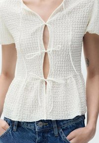Blusa bianca con texture, maniche corte, dotata di scollatura a V profonda e chiusura con stringa in vita, abbinata a pantaloni corti in denim.