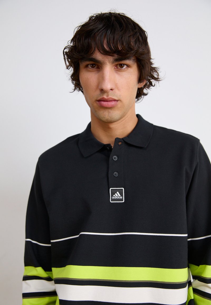 Polo noir avec des rayures horizontales vertes, blanches et noires. Doté d'un col, de trois boutons et d'un logo Adidas sur la poitrine.