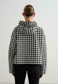 Vrouw met een gevlochten knot draagt een zwart-witte houndstooth-patroon capuchonjas en zwarte leggings, met de rug naar de camera gericht.