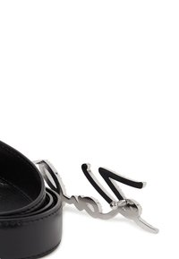 Curea din piele neagră cu o cataramă din metal argintiu lucios, prezentând un design scriptat unic. Catarama are un finisaj lustruit și detalii complexe.