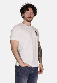 T-shirt bianco a maniche corte con una piccola grafica multicolore sul lato sinistro del petto. Realizzato in cotone, ha una vestibilità rilassata e un collo rotondo.