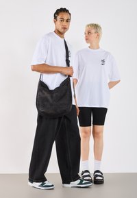A man with braided hair wears a white t-shirt and black jeans, holding a bag. A woman in a white t-shirt and black shorts stands nearby, looking at him.