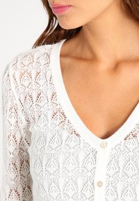 Mujer vistiendo un cárdigan blanco de punto abotonado con un patrón de encaje, mostrando el escote y parte de su rostro y cabello.