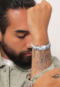 Silverflätat armband med ett lås som har en logotyp; bärs på en tatuerad underarm, vilket framhäver dess texturerade design.
