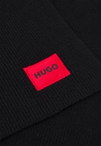 Sort ribbet stof med en rød rektangulær lap, der har teksten "HUGO" i sort på en blød tekstilbaggrund.