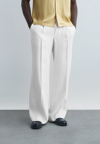 Homme portant un pantalon blanc ample plissé, des chaussures noires et une chemise beige à manches courtes, les mains dans les poches, sur un fond neutre.