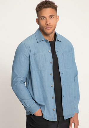 Lichtblauw spijkerhemd met lange mouwen, knoopsluiting, twee borstzakken en een puntkraag, gedragen over een zwart shirt.