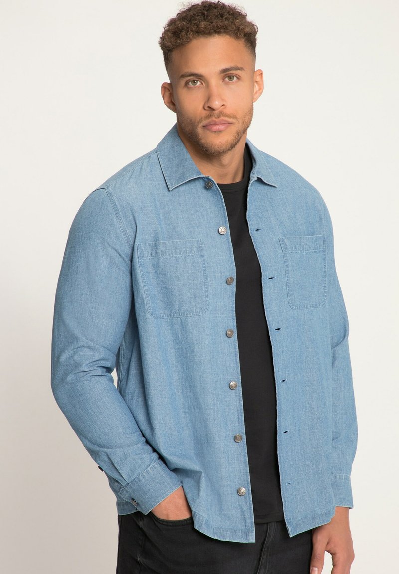 Lichtblauw spijkerhemd met lange mouwen, knoopsluiting, twee borstzakken en een puntkraag, gedragen over een zwart shirt.