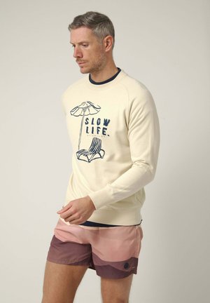 Hombre con jersey crema con el gráfico "Slow Life" y una silla de playa, combinado con pantalones cortos de bloques de color rosa y granate, sobre fondo liso.