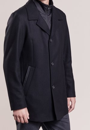 Schwarzer Wollmantel mit strukturiertem Kragen, vier Knöpfen, Seitentaschen und glattem Innenfutter. Schlankes Design mit maßgeschneiderter Passform.
