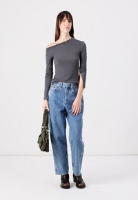 Parte superior gris de manga larga y un hombro descubierto, jeans de mezclilla azul de corte relajado y botas negras hasta el tobillo. Modelo sosteniendo un bolso verde. Fondo simple.