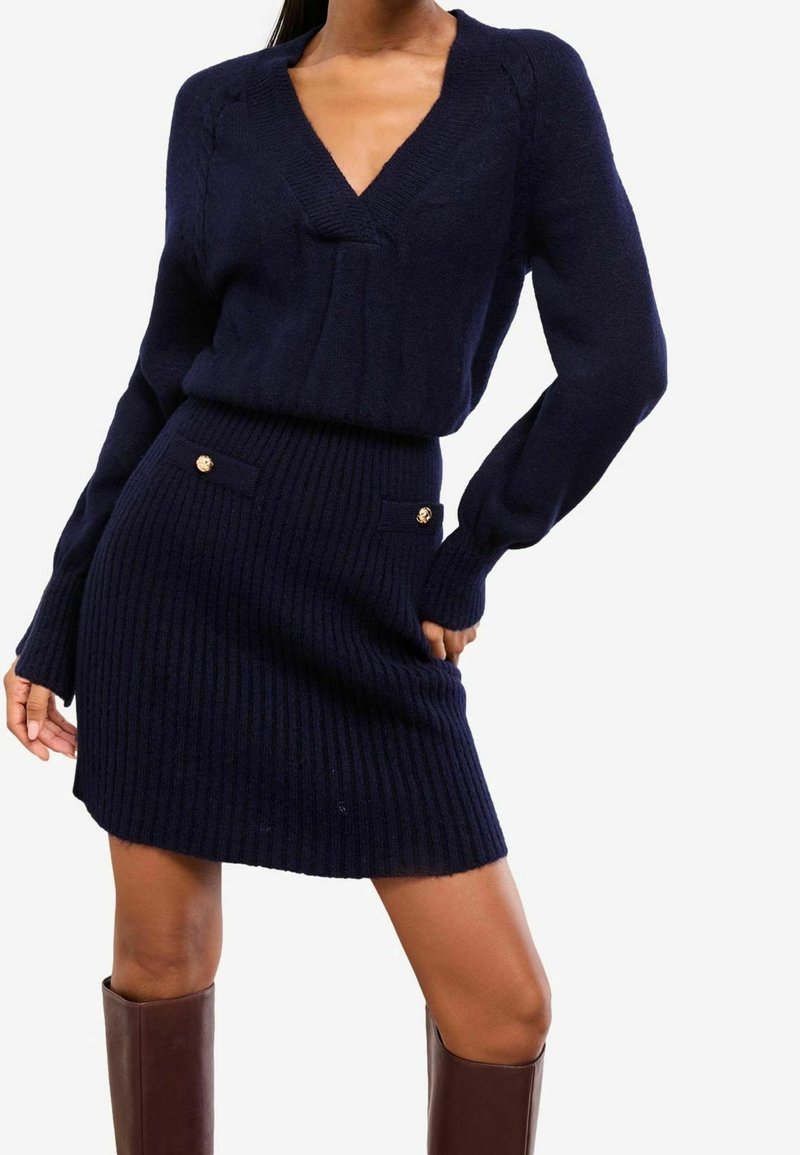 Femme portant un pull en maille côtelée bleu marine à col en V et une jupe assortie avec des détails à boutons dorés, associée à des bottes marron montantes jusqu'au genou.