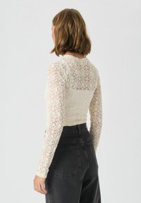 Top din dantelă cu mânecă lungă, de culoare crem deschis, cu un model floral; design ajustat; mâneci透透; asortat cu blugi negri cu talie înaltă.