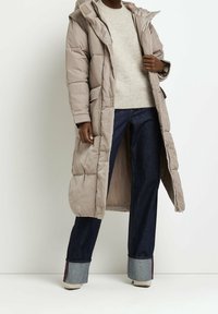 Abrigo largo de plumas beige con capucha, que presenta una textura acolchada y bolsillos frontales, usado sobre un suéter ligero y jeans oscuros.