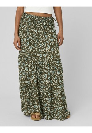 Femme portant une longue jupe fleurie vert olive avec une taille smockée et des sandales beige, debout devant un fond uni.