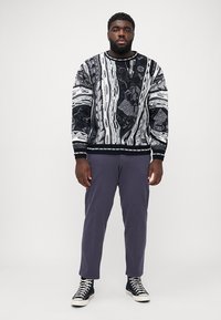 Black and white patterned sweater with long sleeves, crew neck, and ribbed cuffs, paired with purple pants and high-top black sneakers.