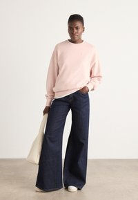 Lyserosa genser, blå vide jeans, hvite sneakers, holder en kremfarget håndveske. Glatt stoff, uformell design, avslappet passform.