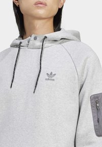 Šedá bavlněná mikina s kapucí, má zipovou kapsu na rukávu, nastavitelné stahovací šňůrky na kapuci a vyšívané logo Adidas na hrudi.