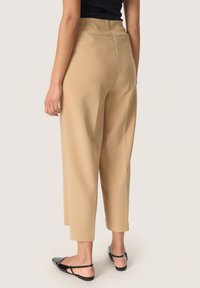 Pantaloni beige a vita alta con gamba dritta e tasche laterali, caratterizzati da una consistenza liscia e una leggera piega sul retro. Scarpe nere basse.