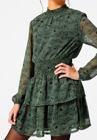 Zielona, przejrzysta sukienka z marszczonym pasem, długimi rękawami, warstwowymi falbanami oraz czarnym wzorem kosmicznym przedstawiającym gwiazdy i planety.