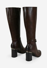 Un par de botas altas de cuero marrón oscuro con tacones cuadráticos y cremallera lateral, que presentan costuras sutiles y una pequeña hebilla cerca del tobillo.
