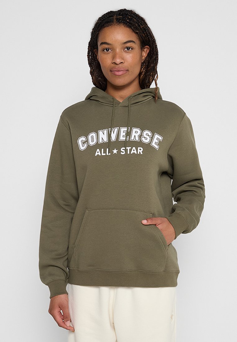Person in olivgrüner Converse All Star Kapuzenpullover mit Fronttasche und cremefarbener Hose, stehend vor einem einfarbigen Hintergrund.