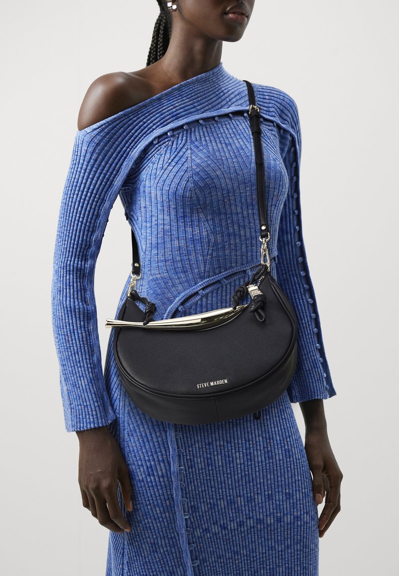 Femme portant une robe bleue côtelée épaules dénudées et un sac bandoulière noir Steve Madden avec des ferrures dorées.