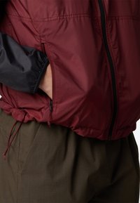Giacca leggera rossa e nera con tasca con zip, orlo regolabile con cordino e una texture liscia e lucida.