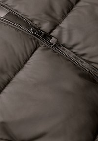 Giacca a vento marrone in tessuto con design trapuntato, caratterizzata da una zip nera prominente e una superficie testurizzata. Dettagli includono una linguetta in tessuto per la zip.