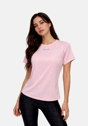 Woman with long dark hair wearing a light pink short-sleeve shirt and black patterned leggings, standing against a plain background.
