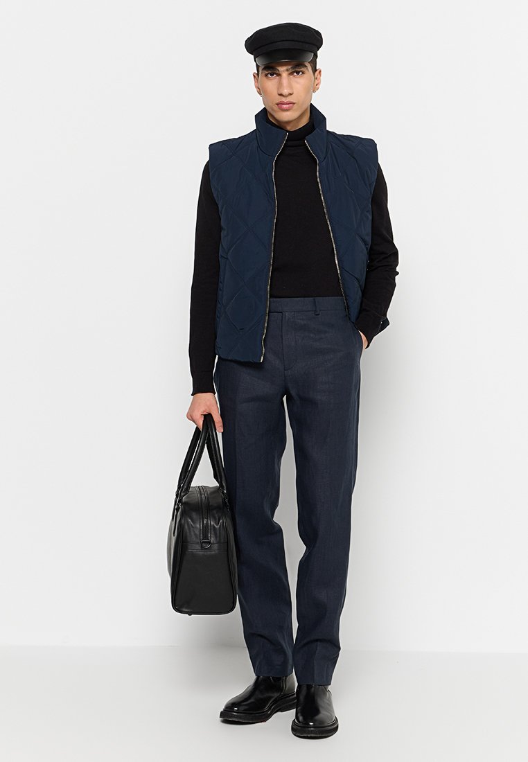 Marineblauer gesteppter Weste über einem schwarzen Rollkragenpullover, kombiniert mit dunklen, maßgeschneiderten Hosen. Das Model trägt eine schwarze Tasche, eine Schirmmütze und elegante Schuhe.