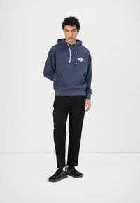Felpa con cappuccio di colore blu navy con lacci bianchi e tasca frontale, caratterizzata da un logo a diamante blu chiaro sul petto, indossata con pantaloni neri.