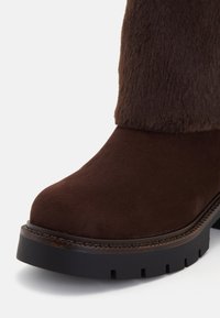 Barna velúr bokacsizma műszőrme felső résszel, kerek orral és vastag fekete gumitalppal, amely vízszintes talpmintázattal rendelkezik.