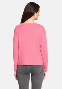 Różowy sweter o luźnym kroju, okrągłym dekolcie i długich rękawach. Materiał wygląda na miękki. Noszony z szarymi dżinsami, prezentuje casualowy styl.
