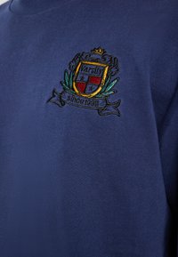 Námořnická modrá mikina se vyšívaným znakem s nápisem "Varsity" a "od roku 1998", ozdobená detaily listů a koruny. Hladká textura.