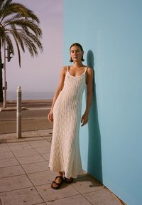 Weißes, strukturiertes Strick-Maxikleid mit dünnen Trägern, getragen mit schwarzen Flip-Flops, vor einer einfarbigen türkisfarbenen Wand und Palmenhintergrund.