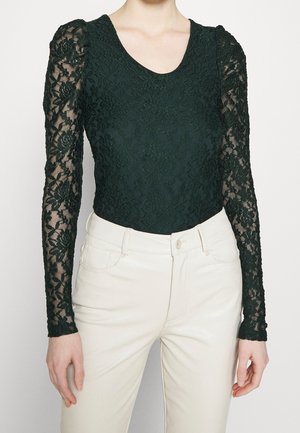 Czarna koronkowa bluzka z długim rękawem ozdobiona kwiatowymi wzorami, zestawiona z białymi spodniami z wysokim stanem, na stojącej osobie.