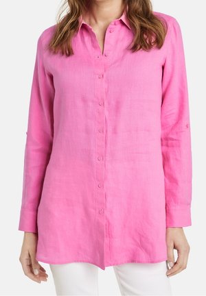 Chemise en lin à manches longues et boutonnée sur le devant, de couleur rose vif, portée par une femme à la peau claire et aux cheveux bruns, style décontracté avec un pantalon blanc.