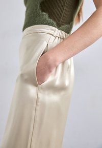 Beezist satin positiivne elastse vöökohtaga, millel on küljetaskud ja sile tekstuur, koos rohelise kudumisega topiga.