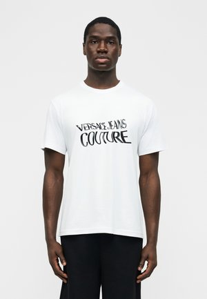 Moški, ki nosi belo majico s kratkimi rokavi z logotipom "Versace Jeans Couture", stoji ob preprostem svetlem ozadju.